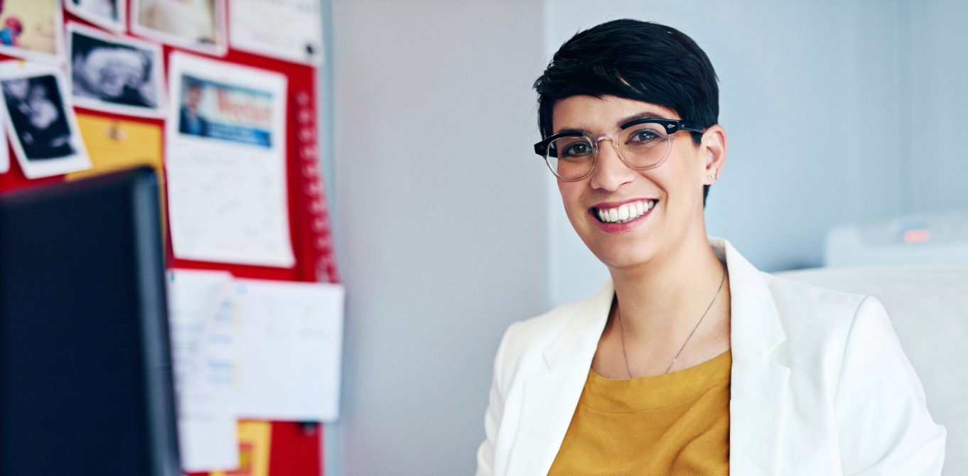 A woman with short dark hair and glasses sits smiling at a desk in an office.
