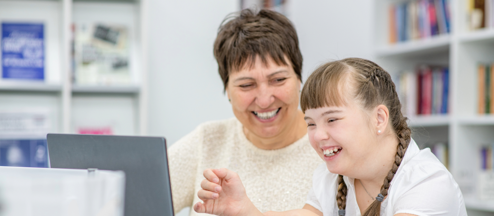 A woman and a young girl smile while looking at a laptop in a library setting.