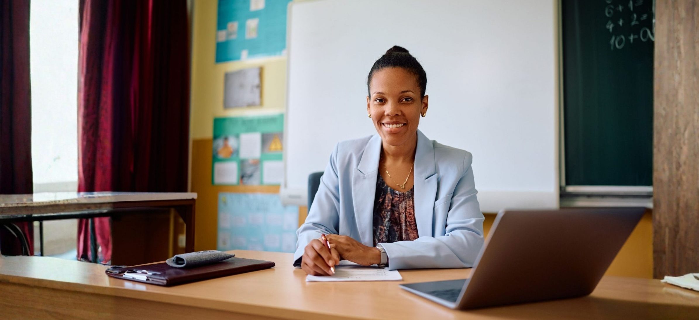 Smiling woman in a light blue blazer sits at a desk in a classroom, with a laptop and notepad.