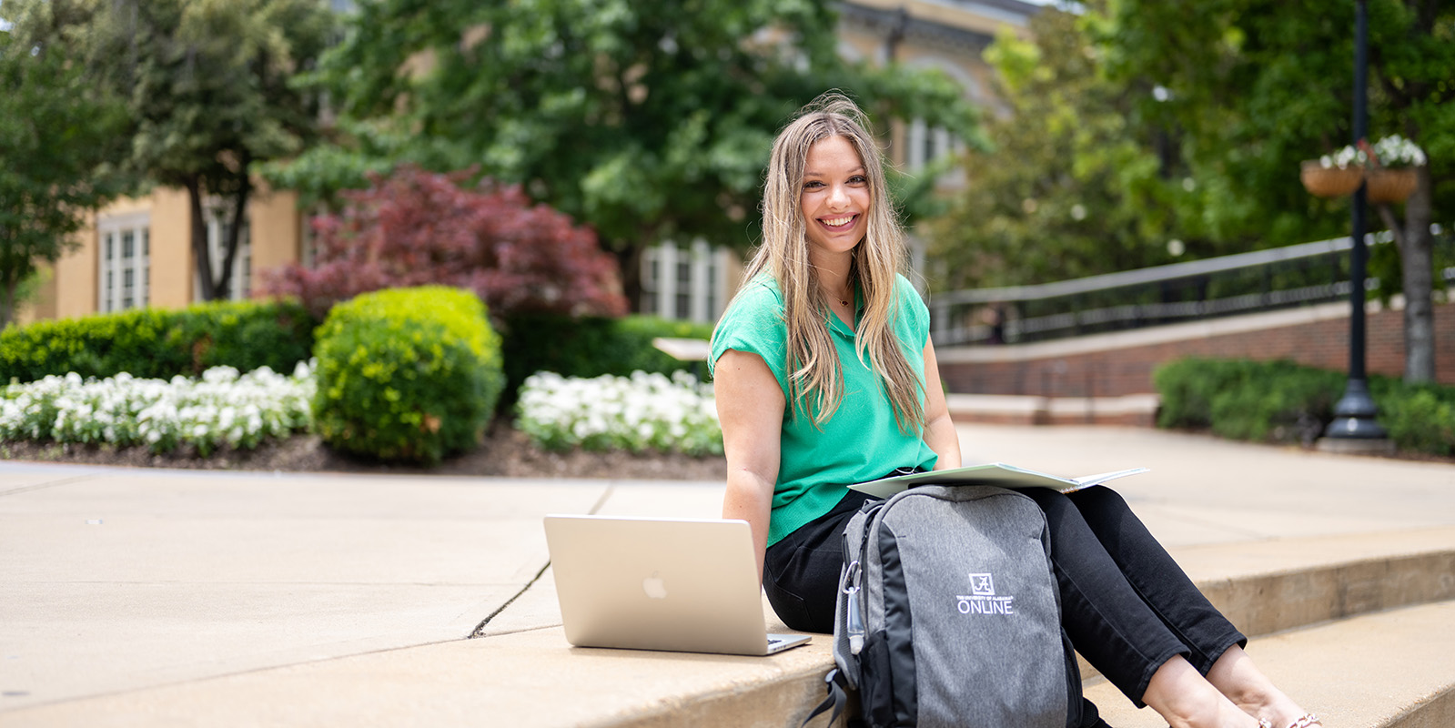 Smiling woman in a green shirt sits on a campus sidewalk with a laptop, notebook, and a backpack.