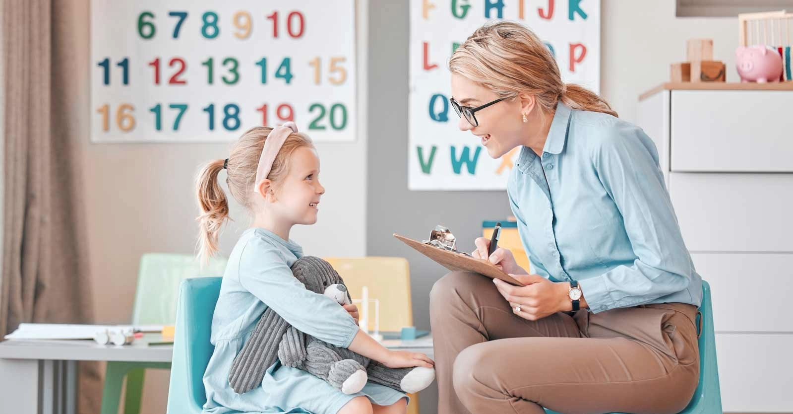 A teacher smiles while talking to a young girl holding a stuffed toy.