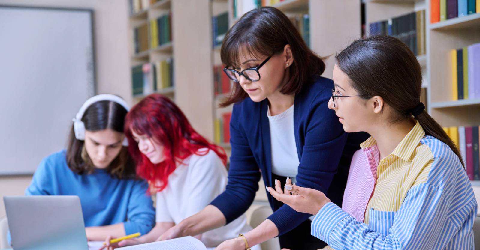 A teacher helps a student with an assignment while classmates study in the background.
