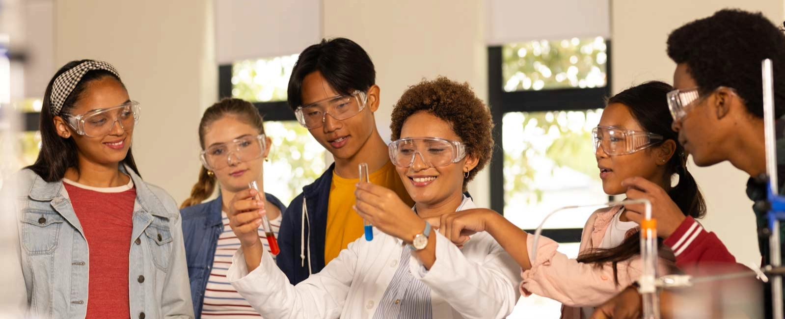 A group of students in a lab watches as a teacher in a white coat and safety goggles holds a test tube.