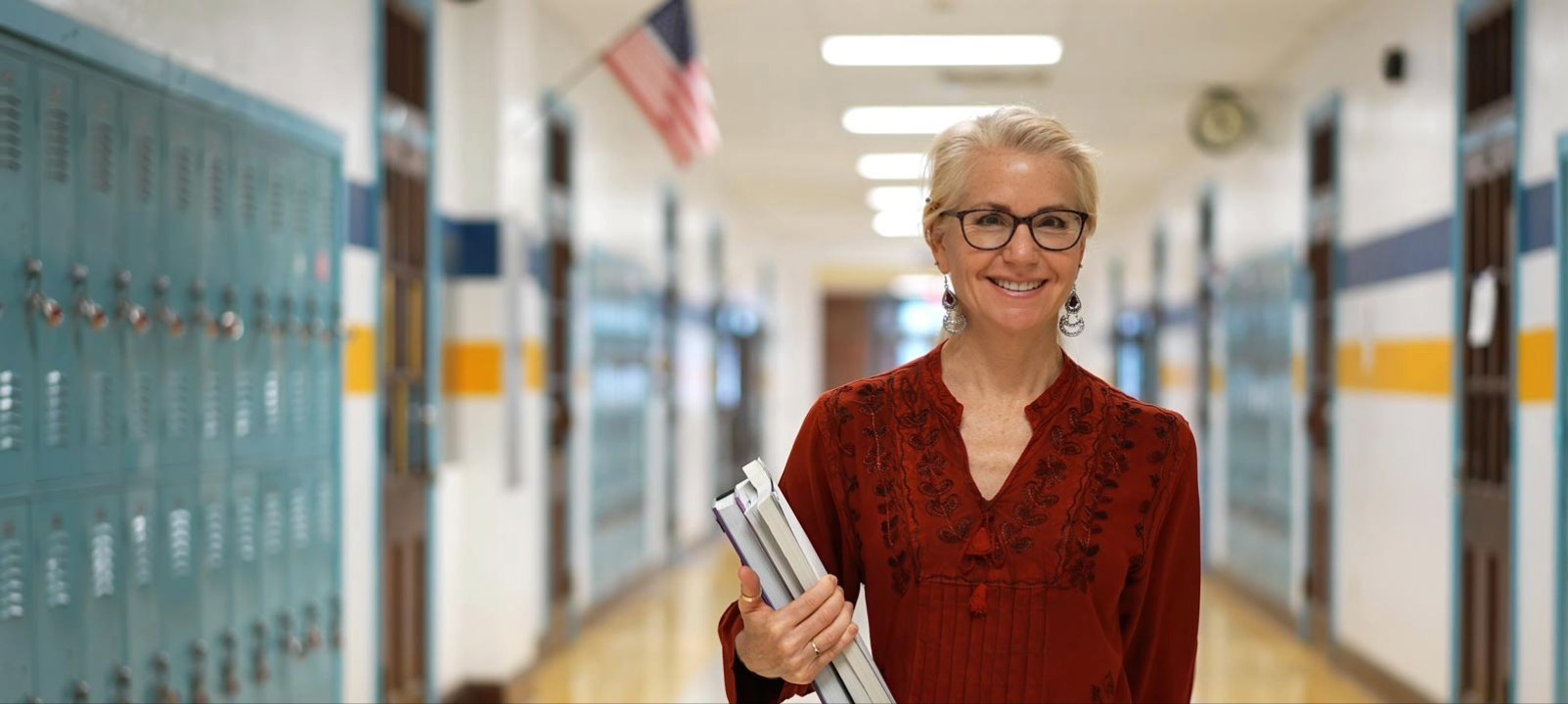 A smiling teacher in glasses stands in a school hallway, holding books.
