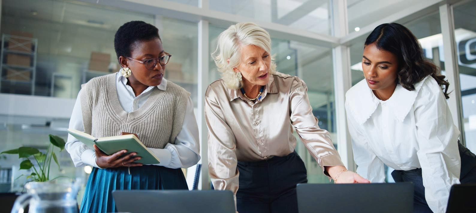 Three women gathered around a table in an office, discussing over laptops and documents.