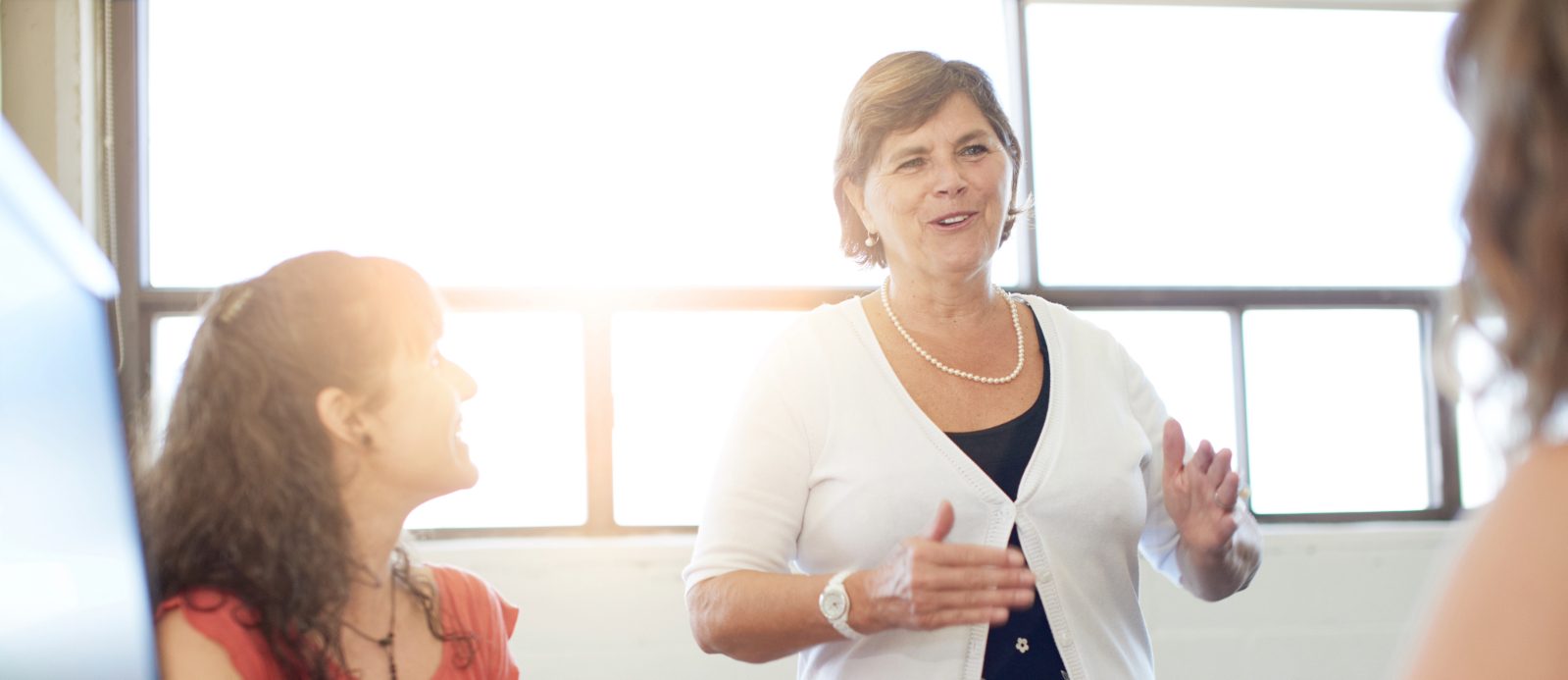 A woman stands and speaks to two seated women in a bright office setting.