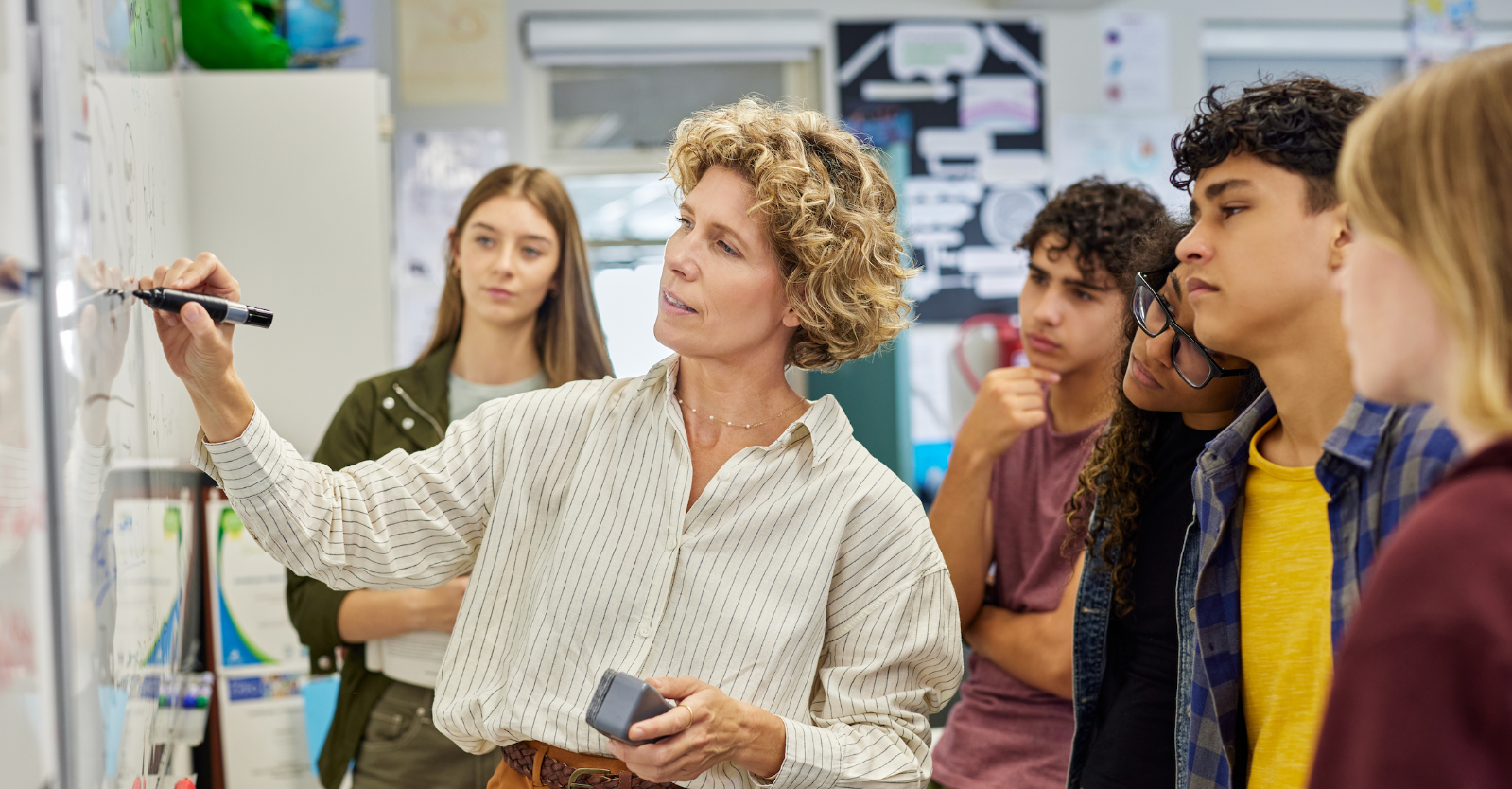 A teacher writes on a whiteboard in a classroom, surrounded by attentive students.