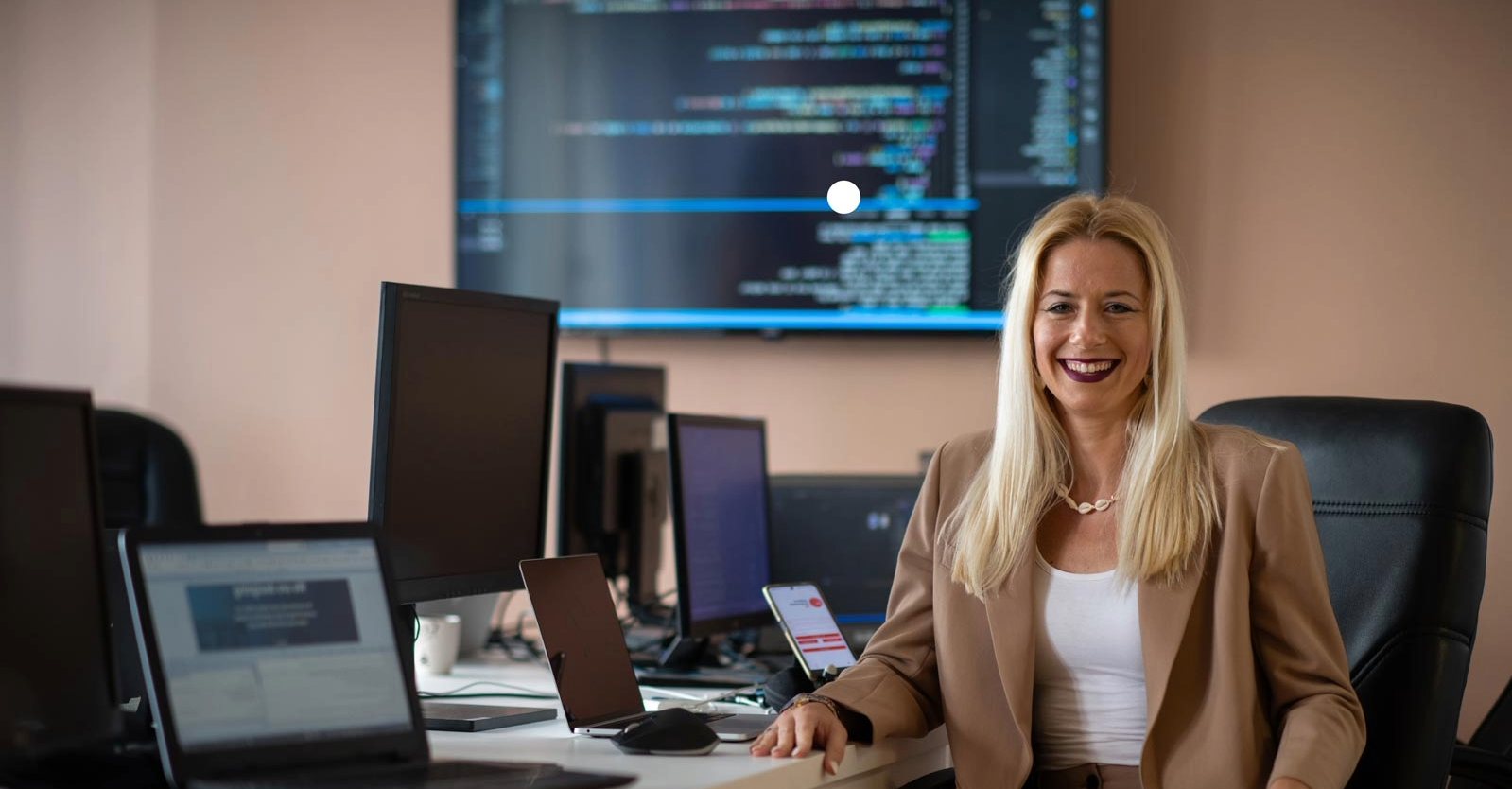 A woman smiles, seated at a desk with multiple monitors displaying code.