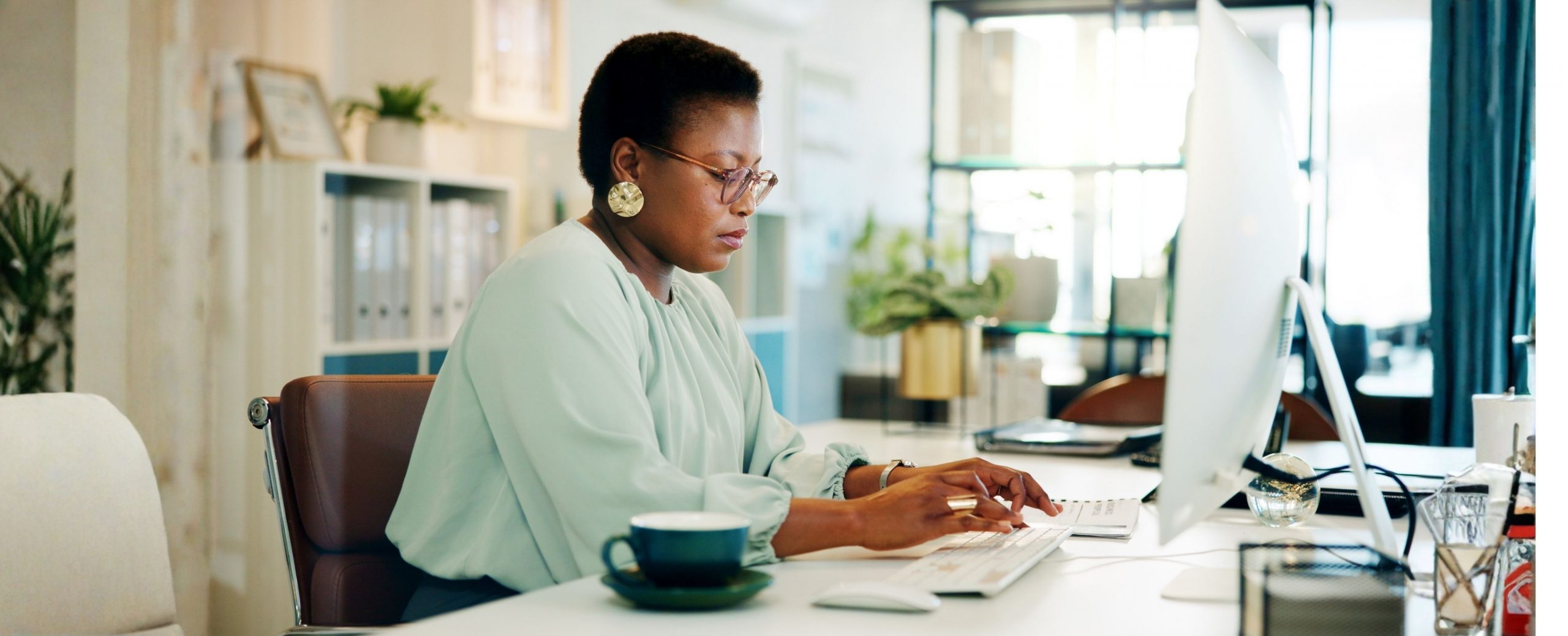 A woman in glasses and a light blouse works at a computer in a bright office.