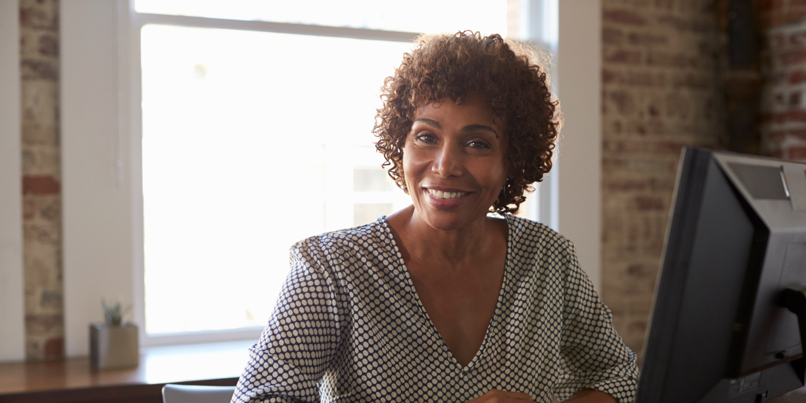 A woman sits at a desk in front of a computer monitor, smiling at the camera in a bright office with a brick wall and a window in the background.
