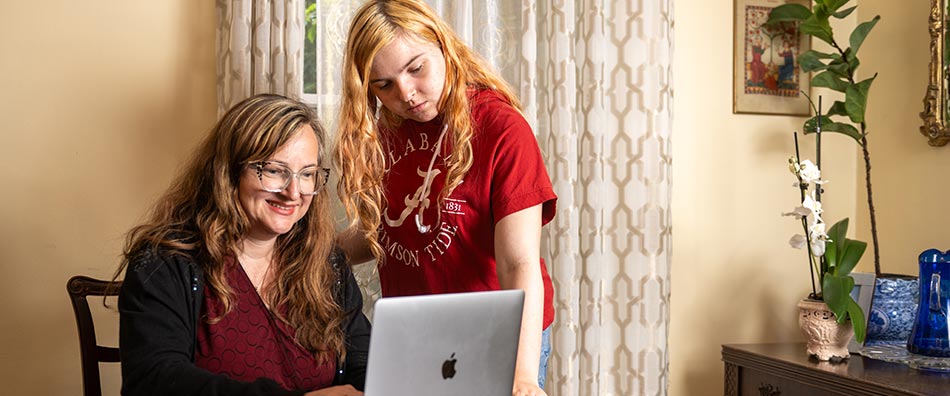 Mother using a laptop sitting at a table with her daughter standing next to her.